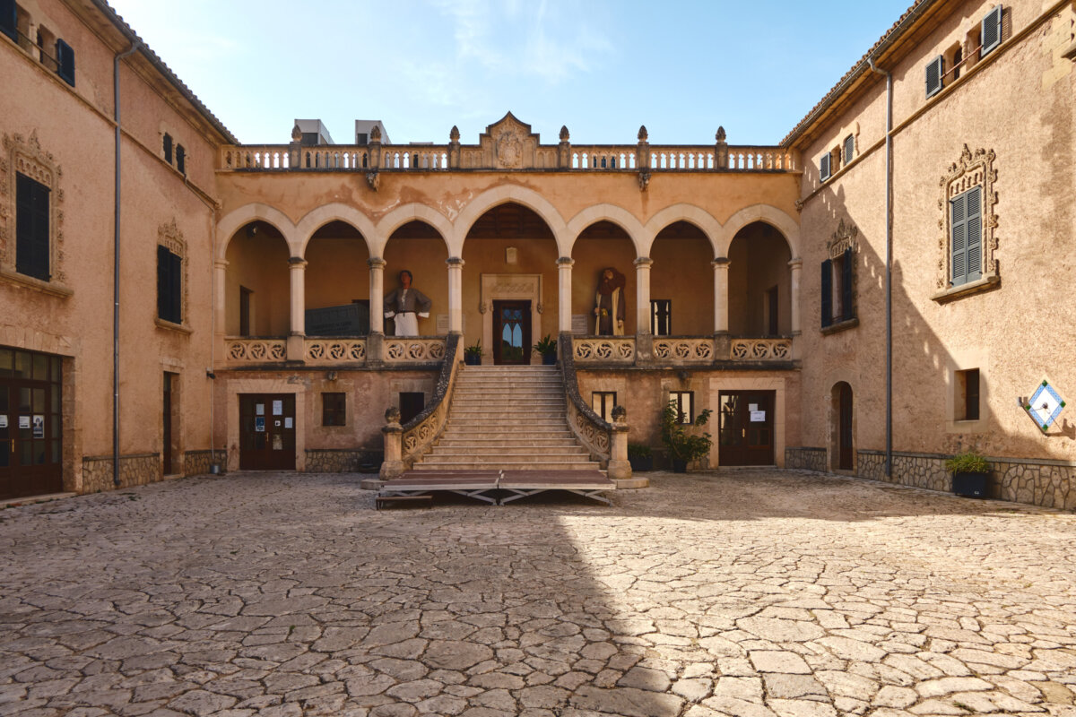 Historischer Innenhof mit Arkaden und Treppen, von Sonnenlicht beleuchtet. Alte Gebäude mit verzierten Fenstern.