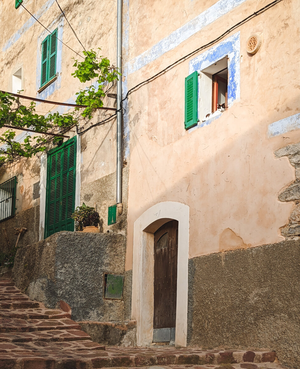 Alter Steinweg vor einem mediterranen Haus mit grünen Fensterläden und einem kleinen Fenster auf der rechten Seite.