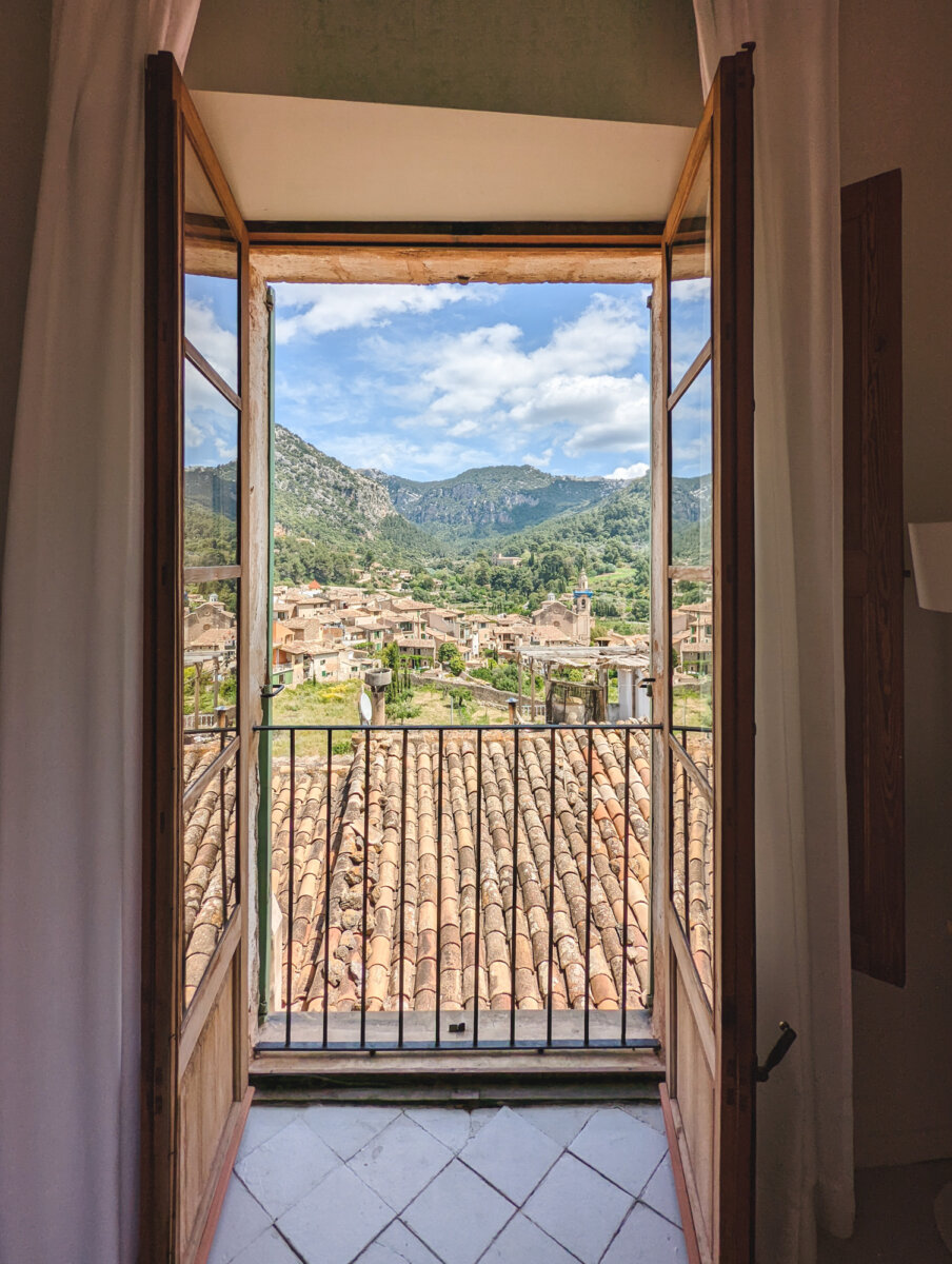 Blick aus einem offenen Fenster auf ein malerisches Dorf mit Bergen im Hintergrund an einem sonnigen Tag.