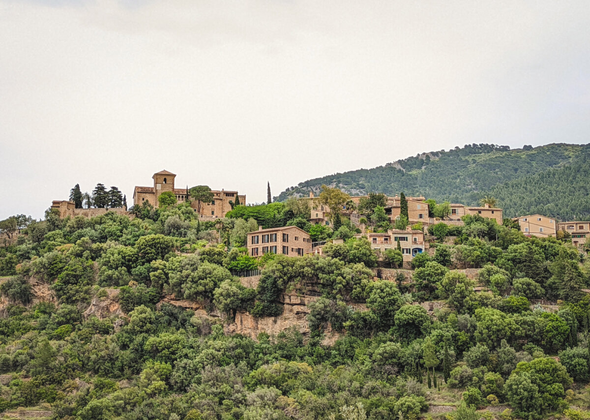 Blick auf ein Dorf auf einem grün bewachsenen Berghang mit alten Steingebäuden und Bäumen.