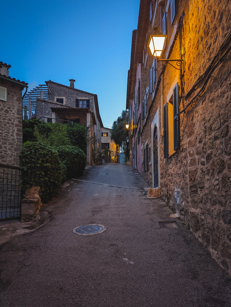 Pflasterstraße in einem mediterranen Dorf bei Dämmerung, mit Straßenlaternen und historischen Steinhäusern.