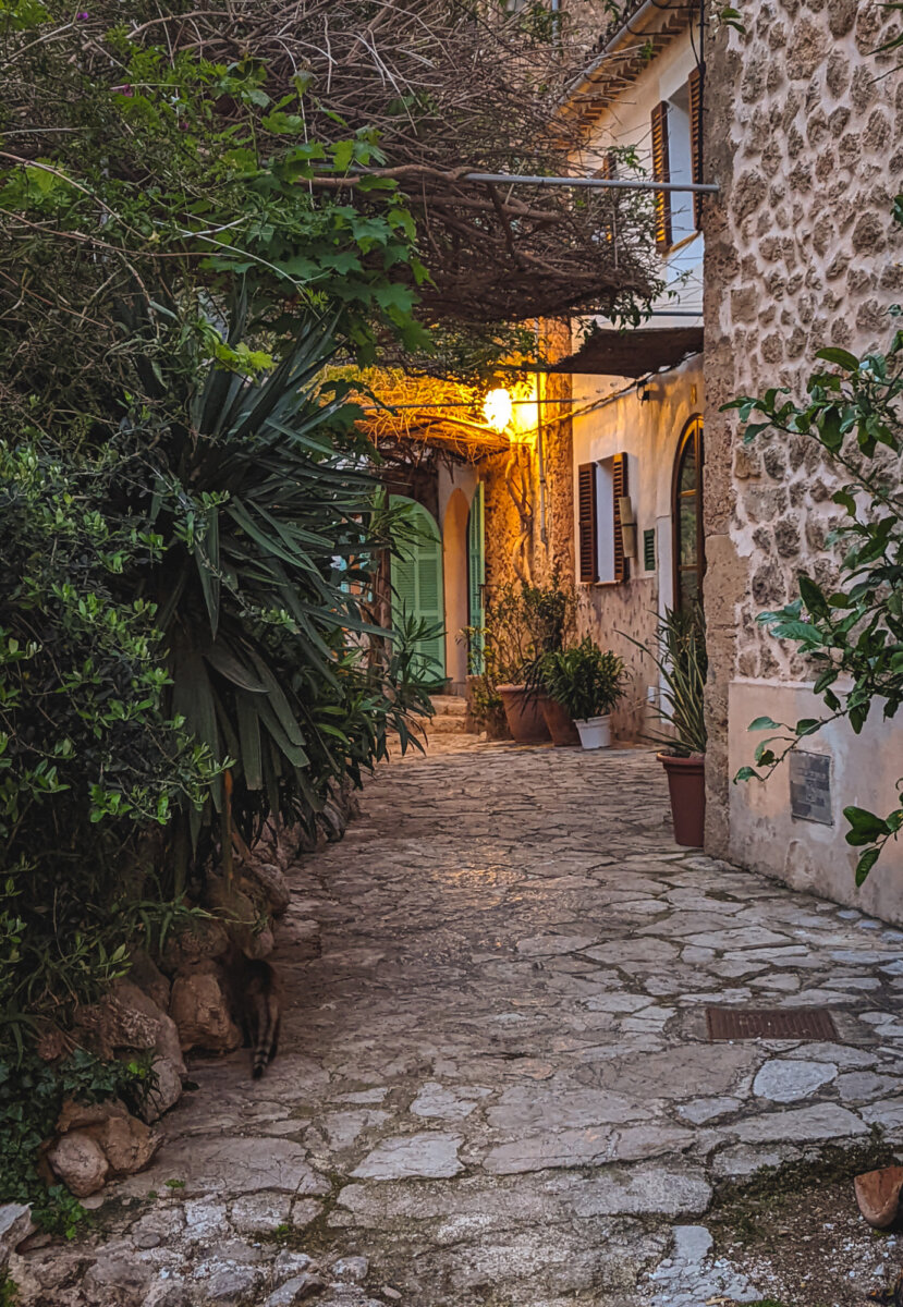 Steinpfad zwischen alten Gebäuden mit Pflanzen und gelber Beleuchtung am Abend in einem mediterranen Dorf.