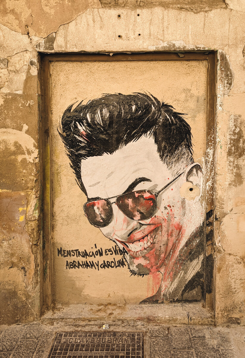 Wandgraffiti einer lächelnden Person mit Sonnenbrille, dazu Text MENSTRUACIÓN ES VIDA und den Namen Abraham y Carolina.