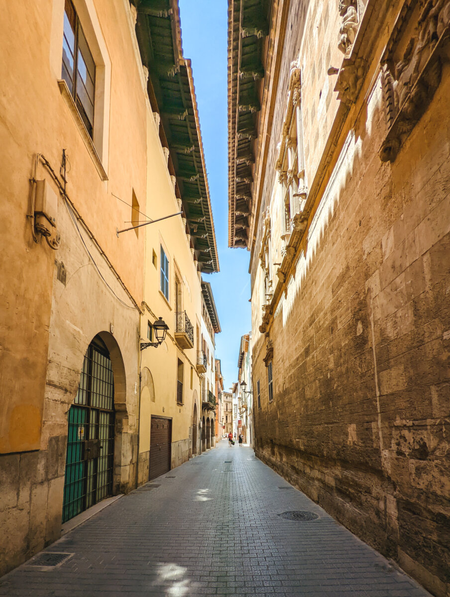 Schmale, sonnige Gasse mit alten, gelben Gebäuden und blauen Himmel in einer historischen Stadt.