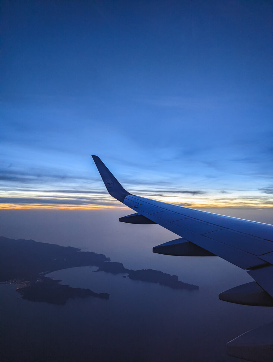 Flugzeugflügel über Inseln in der Dämmerung, Himmel ist in Blau-, Gelb- und Orangetönen gefärbt.